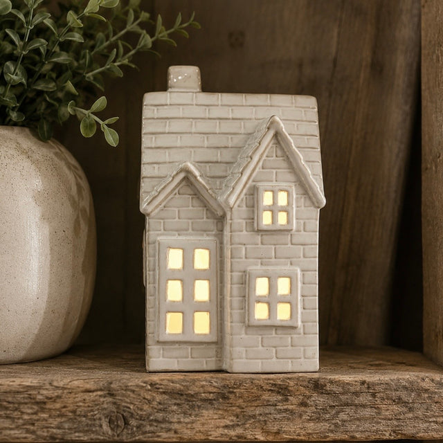 A House shaped ceramic LED tealight with a warm yellow glow from within, displayed on a rustic wooden shelf next to a potted plant.