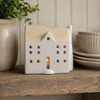 A ceramic house-shaped tea light holder with a warm beige-coloured roof and cream body, shown lit with a small candle inside on a rustic wooden shelf next to a potted plant and stacked plates.