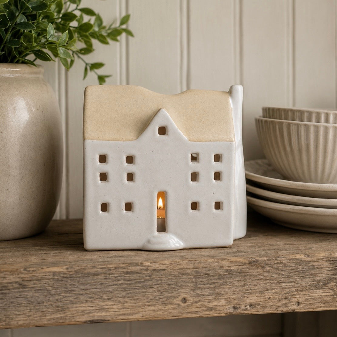 A ceramic house-shaped tea light holder with a warm beige-coloured roof and cream body, shown lit with a small candle inside on a rustic wooden shelf next to a potted plant and stacked plates.