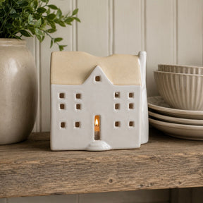 A ceramic house-shaped tea light holder with a warm beige-coloured roof and cream body, shown lit with a small candle inside on a rustic wooden shelf next to a potted plant and stacked plates.