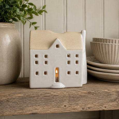 A ceramic house-shaped tea light holder with a warm beige-coloured roof and cream body, shown lit with a small candle inside on a rustic wooden shelf next to a potted plant and stacked plates.