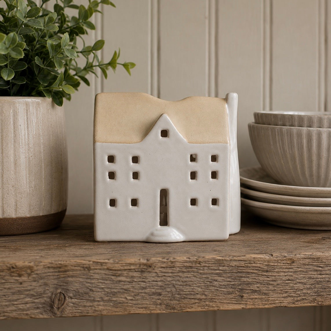 A ceramic house-shaped candle holder or decorative container with a cream body and a warm beige roof, displayed on a rustic wooden shelf next to a potted plant and stacked ceramic bowls.