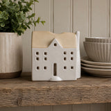 A ceramic house-shaped candle holder or decorative container with a cream body and a warm beige roof, displayed on a rustic wooden shelf next to a potted plant and stacked ceramic bowls.