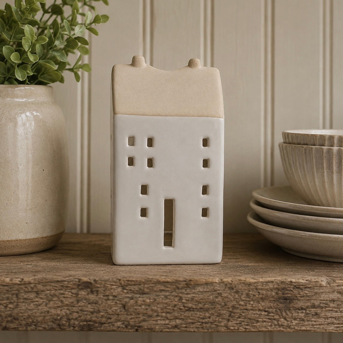 A small ceramic house-shaped candle holder or decor piece in warm cream and off-white, displayed on a rustic wooden shelf with a potted plant and stacked plates in the background.