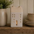 A ceramic house-shaped candle holder with a lit candle inside, displayed on a rustic wooden shelf next to a potted plant and stacked ceramic bowls.