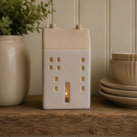 A ceramic house-shaped candle holder with a lit candle inside, displayed on a rustic wooden shelf next to a potted plant and stacked ceramic bowls.