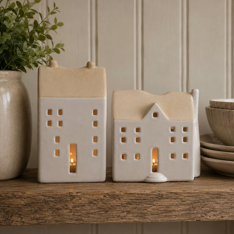 Two small house-shaped ceramic candle holders with lit tea lights, displayed on a wooden shelf next to a vase of greenery and stacked plates.