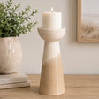 A ceramic candle holder with a tapered base, featuring a glossy off-white upper section and a textured beige lower section, holding a lit white pillar candle on a wooden tabletop.