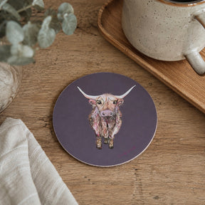 A round coaster featuring a detailed illustration of a highland cow on a dark purple background, styled on a wooden table next to a ceramic mug and a linen cloth.