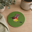A round green coaster featuring a colourful painted hummingbird, styled on a light wood tabletop with a ceramic mug, a folded cloth, and a potted plant.