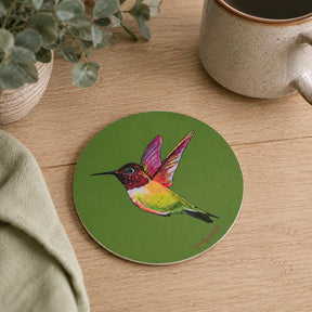 A round green coaster featuring a colourful painted hummingbird, styled on a light wood tabletop with a ceramic mug, a folded cloth, and a potted plant.