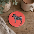A round coral-pink coaster featuring a black and white zebra illustration, styled on a rustic wooden table next to a ceramic mug, a striped cloth, and a potted plant.