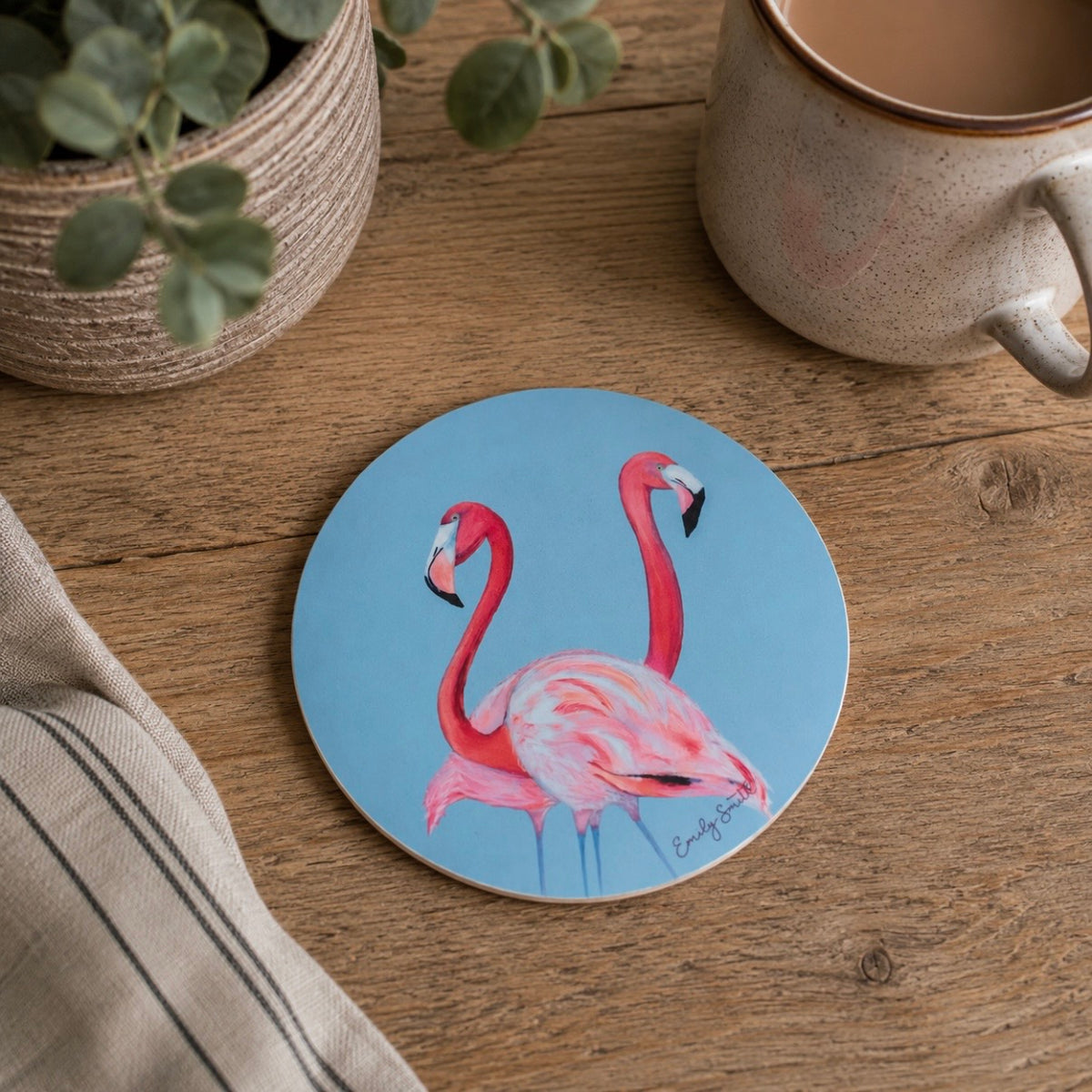 A round coasters featuring a painted illustration of two pink flamingos on a light blue background, displayed on a wooden tabletop next to a ceramic mug and a potted plant.