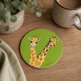 A round green coaster featuring an illustration of two giraffes, styled on a wooden tabletop next to a ceramic mug, a potted plant, and a folded cloth.