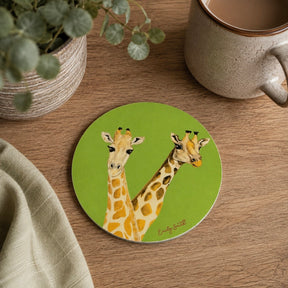 A round green coaster featuring an illustration of two giraffes, styled on a wooden tabletop next to a ceramic mug, a potted plant, and a folded cloth.