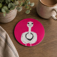 A round pink coaster featuring a white ostrich with black details, styled on a wooden table next to a ceramic mug, a potted plant, and a striped cloth.