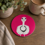 A round pink coaster featuring a white ostrich with black details, styled on a wooden table next to a ceramic mug, a potted plant, and a striped cloth.