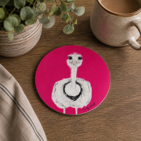 A round pink coaster featuring a white ostrich with black details, styled on a wooden table next to a ceramic mug, a potted plant, and a striped cloth.