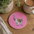 A round pink coaster featuring a painted lemur, styled on a wooden tabletop next to a ceramic mug, a potted plant, and a folded cloth.