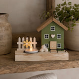 A small decorative wooden house figurine with a green body, natural wood roof and door, white window panes, a picket fence, a flower accent, and a tea light candle on a wooden base, styled on a rustic shelf with a ceramic vase and potted plant in the background.