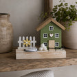 A decorative miniature house figurine with a green body, brown roof, white picket fence, small flower, and tea light candle, displayed on a wooden shelf between ceramic vases.