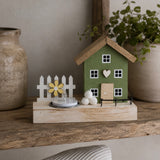 A decorative miniature house figurine with a green body, brown roof, white picket fence, small flower, and tea light candle, displayed on a wooden shelf between ceramic vases.