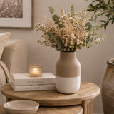 A two-tone ceramic vase with a matte beige top half and white bottom half, filled with dried white flowers and green eucalyptus stems, styled on a round wooden side table in a cozy living room setting.