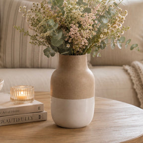 A two-tone ceramic vase with a textured beige top half and a smooth off-white bottom half, filled with dried baby's breath and eucalyptus stems, sitting on a wooden table next to a lit candle and books in a cozy living room setting.