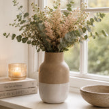 A ceramic vase with a two-tone beige and white design, filled with dried flowers and eucalyptus stems, sitting on a windowsill next to a lit candle, books, and a bowl.