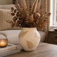 A beige ceramic vase with brown fern leaf patterns, filled with dried pampas grass, brown leaves, and pinecones, styled on a wooden table in a cozy living room setting.