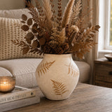 A beige ceramic vase with brown fern leaf patterns, filled with dried pampas grass, brown leaves, and pinecones, styled on a wooden table in a cozy living room setting.