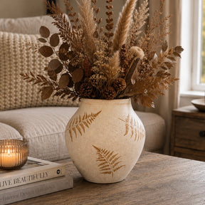 A beige ceramic vase with brown fern leaf patterns, filled with dried pampas grass, brown leaves, and pinecones, styled on a wooden table in a cozy living room setting.