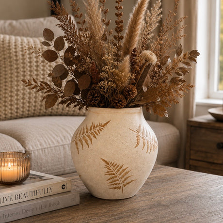 A beige ceramic vase with brown fern leaf patterns, filled with dried pampas grass, brown leaves, and pinecones, styled on a wooden table in a cozy living room setting.