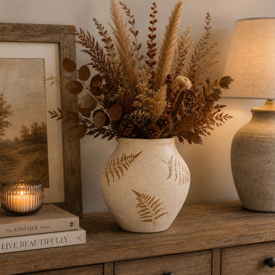 A beige-colored ceramic vase with gold-toned fern leaf patterns, filled with dried pampas grass, brown leaves, and pinecones, styled on a wooden console table next to a lit candle, framed print, and a table lamp.