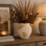 A beige-colored ceramic vase with gold-toned fern leaf patterns, filled with dried pampas grass, brown leaves, and pinecones, styled on a wooden console table next to a lit candle, framed print, and a table lamp.