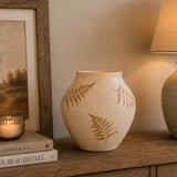 A beige ceramic vase with brown fern leaf patterns, styled on a wooden table next to a lit candle, framed picture, and a lamp.