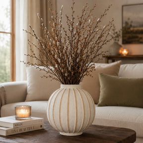 A ribbed cream ceramic vase filled with dried willow branches, styled on a dark wood coffee table in a cozy living room setting.
