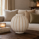 A ribbed ceramic vase in a warm cream color with natural wood stripes, displayed on a dark wooden coffee table in a cozy living room setting.