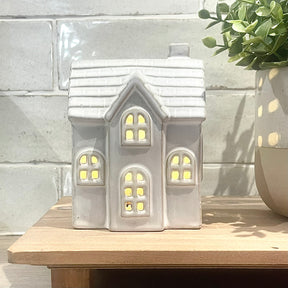 Ceramic house-shaped lantern with glowing interior on a wooden surface against a tiled wall.