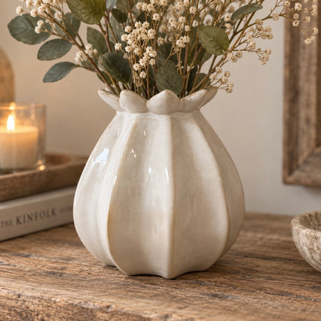 A glossy cream-coloured ceramic vase in a poppy bud shape, holding dried white flowers and green leaves, set on a rustic wooden table next to a lit candle.