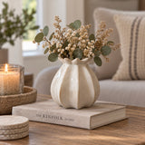 A cream-coloured ceramic vase in a poppy bud shape, filled with dried flowers and green eucalyptus stems, displayed on a beige book on a wooden table in a cozy living room setting.