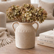 A rustic beige ceramic vase with two handles, filled with dried green and brown hydrangea stems, sits on a wooden table next to stacked books in a cozy living room setting.