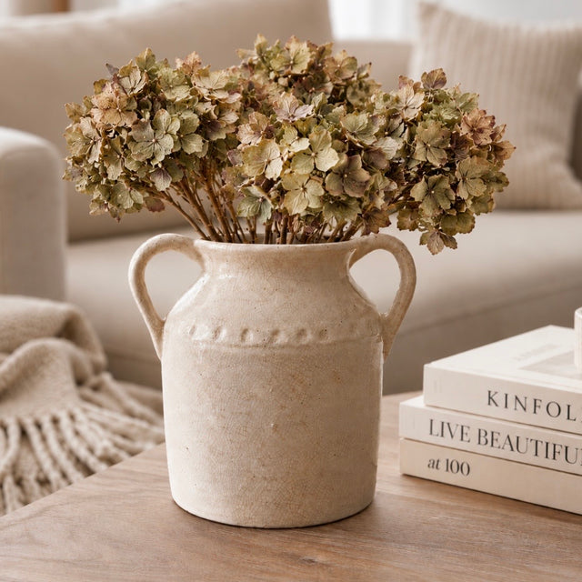 A rustic beige ceramic vase with two handles, filled with dried green and brown hydrangea stems, sits on a wooden table next to stacked books in a cozy living room setting.