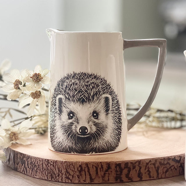 White ceramic pitcher with a hedgehog design on a wooden surface with flowers in the background