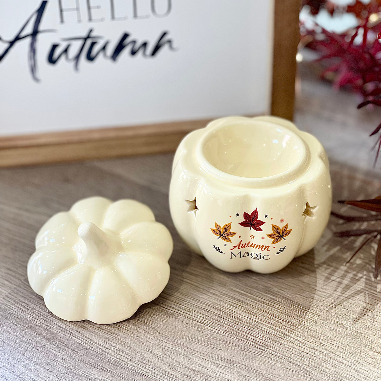 Ceramic pumpkin Wax Burner with 'Autumn Magic' text, placed on a wooden surface and a framed 'Hello Autumn' sign in the background