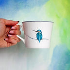 Hand holding a mug with a blue bird design against a colorful watercolor background