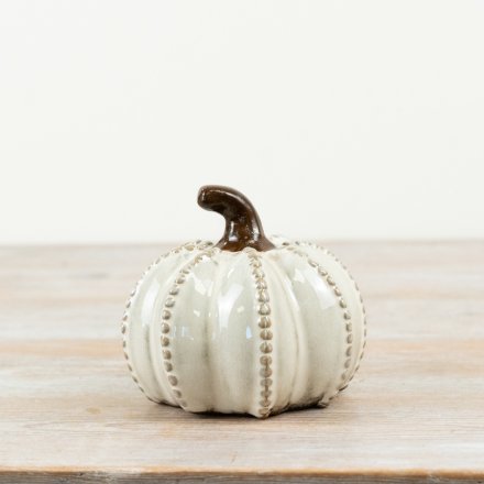 A small decorative ceramic pumpkin in an off-white glaze with a dark brown stem, standing on a light wood surface against a plain light background.