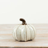 A small decorative ceramic pumpkin in an off-white glaze with a dark brown stem, standing on a light wood surface against a plain light background.