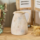 Ceramic pitcher on a wooden surface with a wicker basket and plants in the background