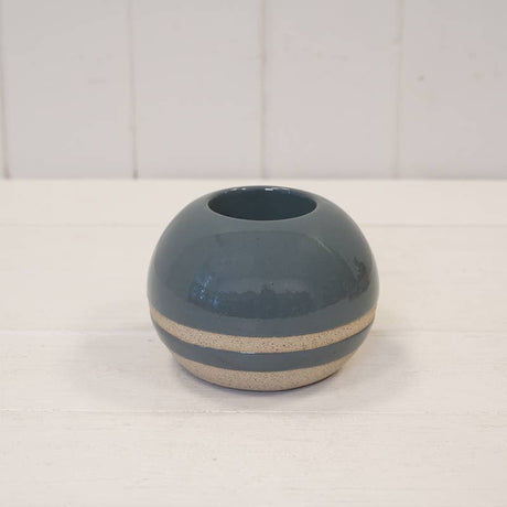 A small round ceramic vase with a glossy dark blue-gray finish and a textured beige band around the middle, set on a white wooden surface against a white paneled background.